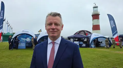Luke Pollard looking at the camera wearing a suit and tie, he is stood on Plymouth Hoe. Tents which have been set up as information points can be seen in the background with Royal Air Force signs.