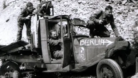 Coleshill Auxiliary Research Team A black and white photo showing four men in Home Guard uniform on a beaten up flat-back lorry, which has the word "Berlin" and an arrow painted on the side of its bonnet. One man is crouched on the bonnet, with a pistol in his right hand; a second is sitting behind the wheel; the third and fourth are on the flat back, one crouched with a pistol and the other pointing a machine gun. 