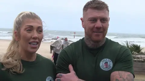 Woman with long blonde hair and man with short fair hair stand together wearing green t-shirts a beach and sea can be seen behind.