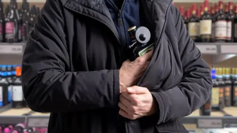 Getty Images A man, who is standing inside a shop, tried to hide a bottle of alcohol with a security tag attached under his dark coloured coat. Behind are shelves full of different bottle of alcohol.