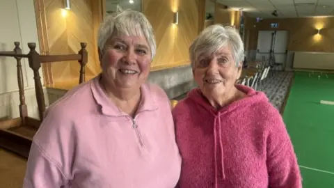 A close-up of Margaret and Barbara standing next to one another and smiling. Both ladies have short grey hair: Margaret, on the left, wears a pale pink top with a half zip, Barbara wears a bright pink, woolly jumper with a hood. The interior of the bowling club can be seen behind them.