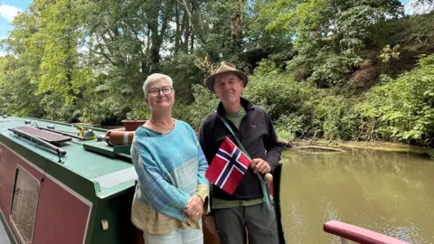 Hugh Pagler and his wife Wendy stand on their canal boat. He is wearing a brown hat and black jumper and holding a Norwegian flag. Wendy is wearing a blue and yellow striped jumper and has short white hair and glasses. Behind them is the canal.