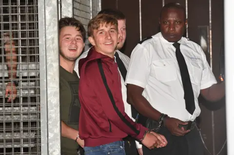 PA Albert Bowers (left) and Jessie Cole (centre), smiling, appearing to laugh or mock the assembled media, as they leave Reading Magistrates Court following an appearance in relation to the death of PC Andrew Harper. 