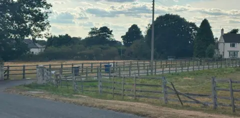 LDRS Two fields in a rural village, surrounded by trees and wooden fencing. A white house with a red roof is on the right of the picture