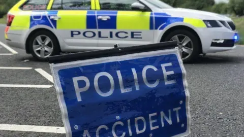 A blue sign saying police accident in white capital letters. It is sitting on a tarmacked road. Behind it is a white, blue and yellow police car and behind that, a verge. 