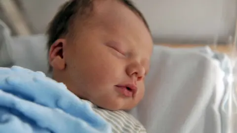 A baby sleeping in a cot. It has dark hair and is covered by a blue blanket. 
