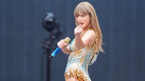 Taylor Swift performing in a sparkling gold and blue leotard, holding a blue and gold microphone and looking at the camera