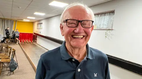 Terry is smiling at the camera. He is wearing dark-framed glasses and a navy t-shirt. There is a skittle alley behind him and two people stand in the background.