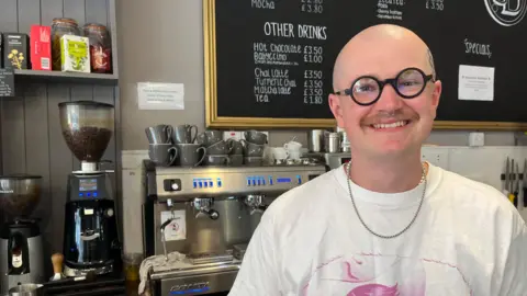 Alfie Tipping stands in front of the coffee machine in Southville Deli and smiles at the camera