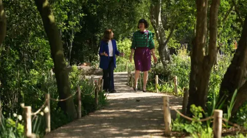 Two women are walking together along a winding, sun-dappled gravel path through a lush woodland area. The woman on the left wears a blue blazer and is gesturing while speaking, while the woman on the right is dressed in a green patterned top and purple skirt.