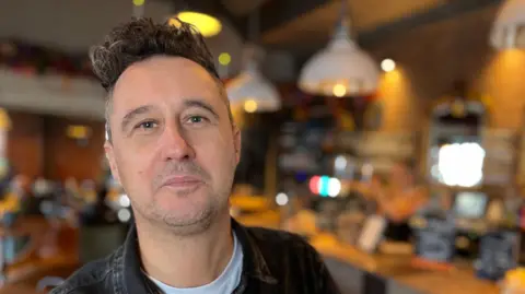 Middle aged man with a dark jacket and white T shirt inside a bar