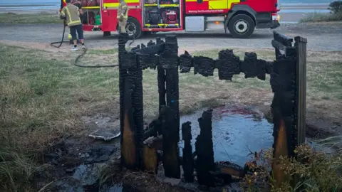 The remains of a cremated bin at Alnmouth beach. The charred wooden structure is almost completely destroyed and there is a puddle of water on the ground around it. In the background are two firefighters carrying hoses attached to a fire engine, which is parked on the seafront.