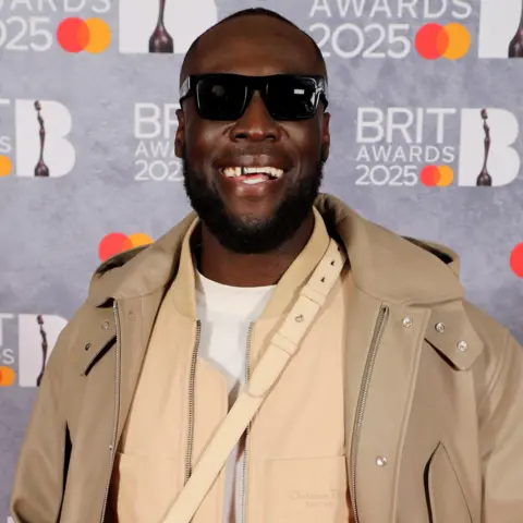 Getty Images Stormzy tersenyum untuk kamera sambil mengenakan kacamata hitam, saat ia berpose di karpet merah Brit Awards.