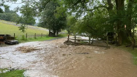 Muddy flood water flowing around a wooden fence and past fields and trees in All Stretton