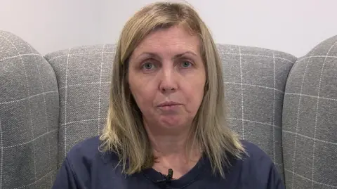 A woman with shoulder-length blonde hair sits on a grey armchair. A white wall is behind the chair. She is wearing a navy blue top.