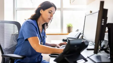 Nurse with long brown hair and blue scrub outfit works at a computer
