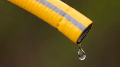 A yellow hose with grey stripes pointing diagonally downwards with a drip of water coming out the end