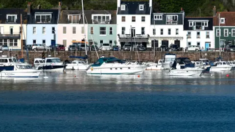BBC Some 14 small yachts and motor boats are moored along a waterfront marked with a harbour wall that has pastel coloured houses behind them.