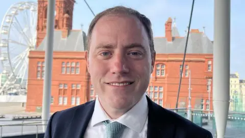 A portrait photograph of Rhys ab Owen, wearing a suit and tie, standing on the Senedd steps with a view of the Pierhead building in Cardiff Bay in the background.