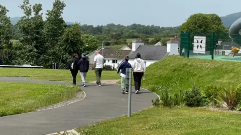 Five people are walking away down a curved walkway with grass either side and trees and houses in the distance.