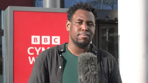A reporter standing with a microphone in front of a red and white BBC cymru wales sign. He has short black hair and thin beard. He is wearing an black jacket open to a green t shirt