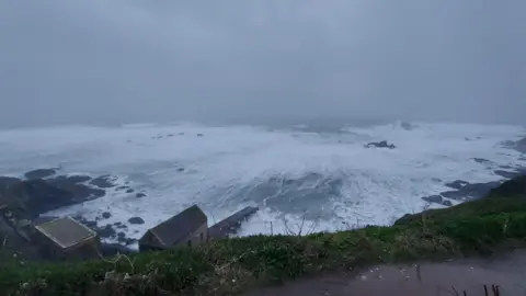 A series of waves are seen rolling into the shore. The image is taken from above looking down. Buildings can be seen on the shore line