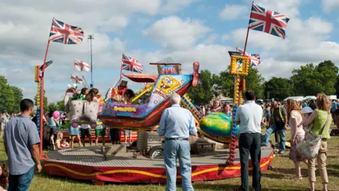 Adults waiting for children having a ride on a brightly coloured, modern merry-go-round in a field. There are other stalls and rides in the background and union jack and Saint George's Cross flags on poles. 