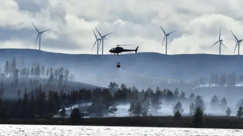 a helicopter dropping water over soking fields with wind turbines on hills in the background