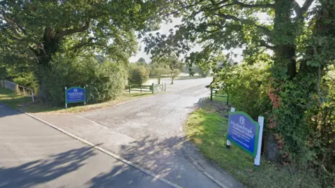 A Google street view screenshot of the entrance of the school. It's a gated entrance on a rural lane with a sign on either side of the road saying the name of the school. No buildings can be seen in the picture, with just a road, a stretch of grassland and some trees visible beyond the open gate.