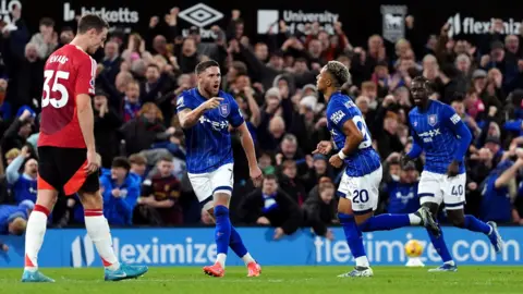 PA Media Ipswich Town players Wes Burns and Axel Tuanzebe run to congratulate Omari Hutchinson after he scored an equaliser to make it 1-1 in the game with Manchester United at Portman Road. Manchester United defender Jonny Evans is also in the picture looking dejected while fans celebrate in the background