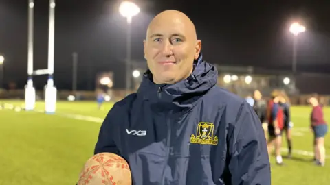 Jacob Kelly smiles at the camera and carrying a rugby ball. He's wearing a navy jacket with the Dings Crusaders RFC logo on the right side. Behind him is a blurred image of a rugby game.