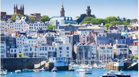 BBC The town of St. Peter Port in Guernsey. In the foreground are boats on the water and behind them is the town, with colorful buildings stacked on a hill.