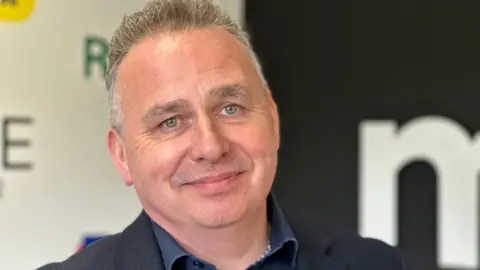 Matthew Marriott pictured smiling. He has short but spiked grey hair, wears a blue blazer and blue collar shirt.