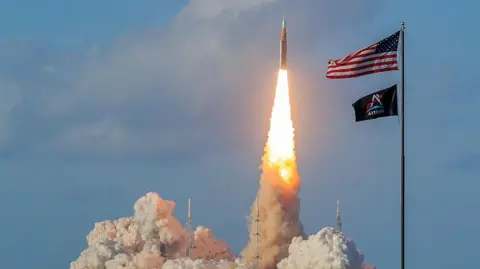 NASA's Artemis II Space Launch System rocket and Orion spacecraft blasts off from the Kennedy Space Center. A vertical pillar of flame erupts from the rocket as it shoots off into the blue sky.
