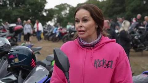 A woman stood in a grass field surrounded by motorcycles. She has long, brown hair tied back and is wearing a bright pink hoodie.