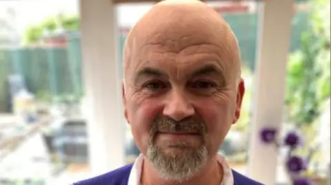 A man stands in a room with glass walls. He has a bald head and a grey beard. He is wearing a purple zip-up t-shirt with a white collar. 