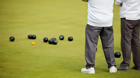 A shot of two people wearing light tops and grey trousers standing on a bowls pitch, you can't see their heads. There are bowls ahead of them on the grass.