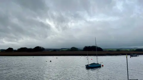 A yawl floating on the Exe estuary close to Topsham with an area of reeds in the background and several buoys visible on the water.