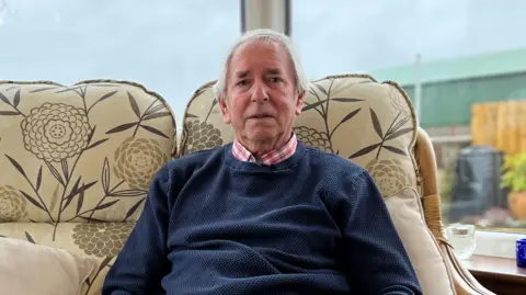 Robert Cooper/BBC Dave Westren looking into the camera. He has short, white hair and is wearing a blue jumper over a pink and white checked shirt. He is sitting on a beige, patterned sofa with flowers on it. It is in front of a large window which looks out into a garden and there looks to be a barn on the right.
