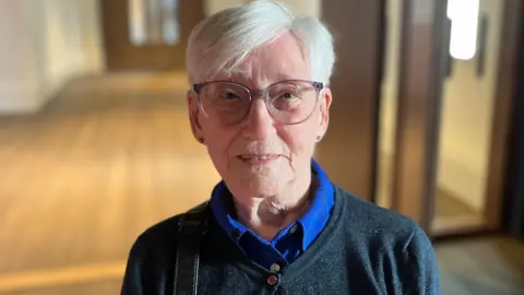 Close-up portrait of 90-year-old Mary with short white hair and glasses, wearing a dark sweater over a blue collared shirt, standing indoors with a softly blurred hallway background.
