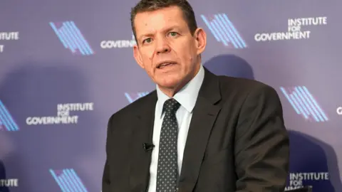 PA Media Rhun ap Iorwerth looking down from a seated position. He is wearing a dark suit and tie, and a white shirt, and is sat in front of a banner for the Institute for Government.
