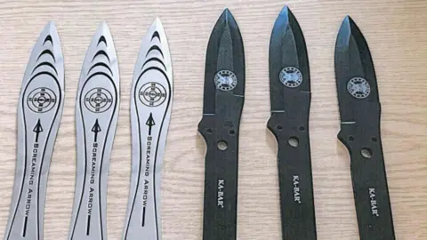 A close-up image shows six throwing knives arranged side by side on a light wooden surface. On the left, three silver-coloured knives are aligned with pointed tips facing upward, each featuring a target-style circular design with curved lines and the text “SCREAMING ARROW” printed along the blade. On the right, three black knives of a similar shape are positioned in the same orientation, each displaying a circular logo near the blade and the brand name “KA-BAR” printed vertically on the handle. All six knives have symmetrical, tapered blades and cut-out holes near the base of the handles, suggesting they are designed for balance and throwing.
