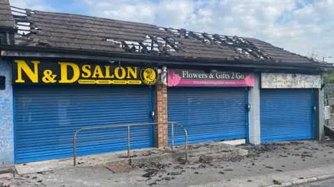 Front view of the shows, all with shutters down and the roof damaged. In places it has almost entirely fallen through.