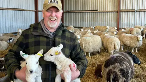 Jim Campbell Jim Campbell is a man in his 60s wearing a warm checked jacket and green hat iwth a goatie beard. He is standing in a barn holding a pair of lambs with a number of ewes behind him 