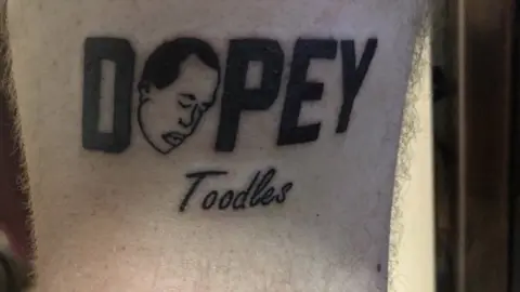 Dopey podcast  A Dopey logo tattoo, with the word 'toodles', on an unknown person's arm