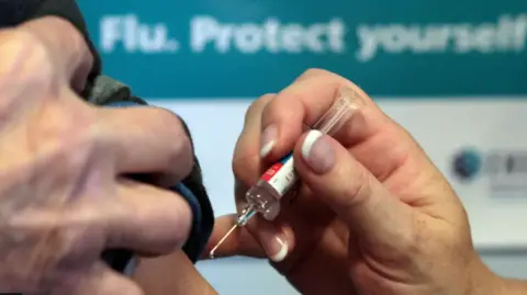 PA Media A close up of a nurse's hand with French manicured nails holding a syringe poised to vaccinate a patient's shoulder. A blurred sign on the wall behind says Flu, Protect Yourself. 