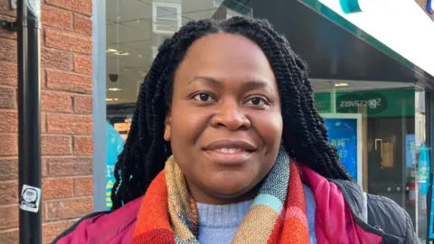 Marthilda Odeode. It is a head and shoulders shot of her. She has long black braids and is smiling at the camera, wearing a light blue jumper, black coat and colourful scarf. 