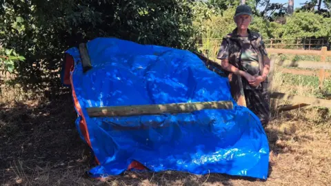 Carol Hurrell A blue sheet covering something large with logs weighing it down. A woman in camouflage is standing next to it.
