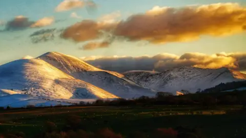 Ian Bell The snow-covered slopes of Skiddaw are lit up in orange by the sun. Clouds have gathered above the fells. There is no snow at the bottom of the mountains.