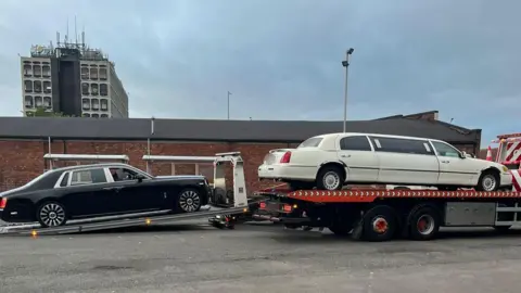 Two luxury cars on the back of a lorry. They are on a car park and made a lengthy convoy. There is little else in the picture apart from a brick building in the background and a grey tower block.